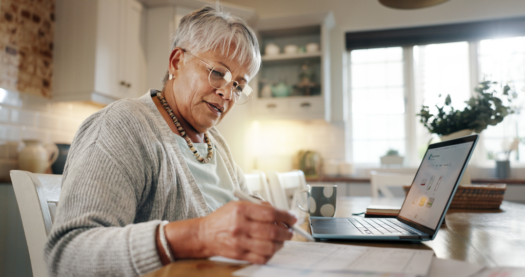Senior woman, sitting in living room with laptop and notebook, researching.