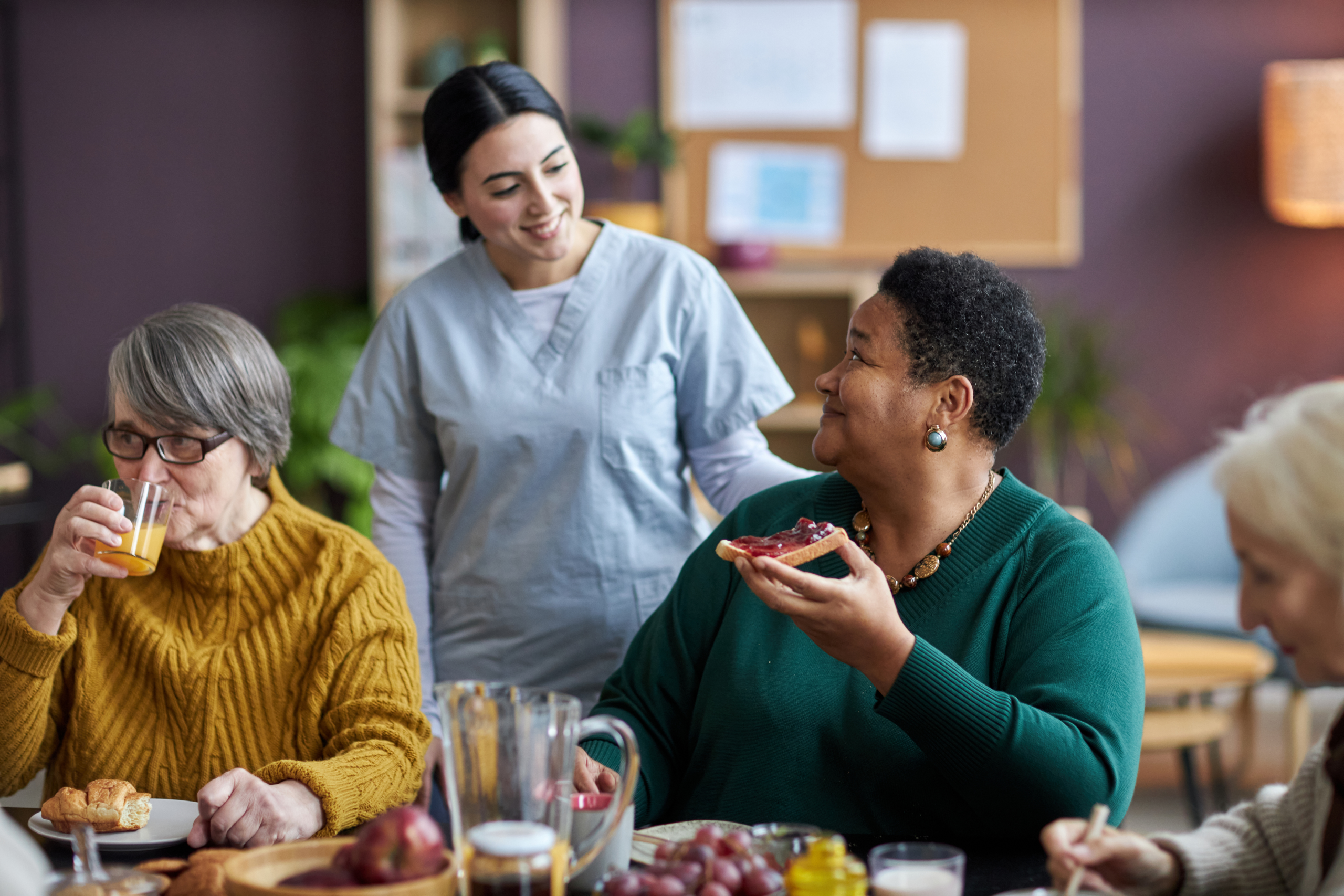 Two seniors sitting down at table for breakfast in senior living community, caregiver/nurse behind them, checking on them and smiling.