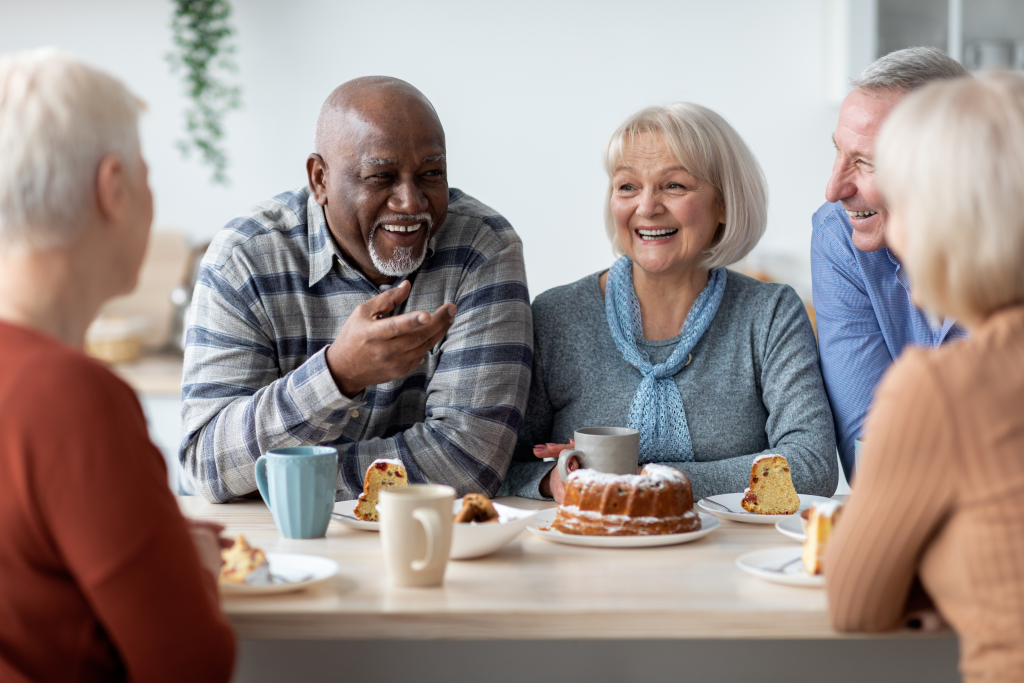 Group of seniors in assisted living room, sitting around table, eating and smiling.