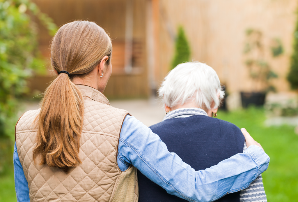 Shot from behind of adult female walking with senior mother on a beautiful, sunny day at senior living community.